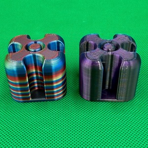 May include: Two 3D printed square-shaped objects with rounded corners and a central circular opening. One object features horizontal stripes of red, blue, and yellow, while the other has a dark, iridescent purple and blue finish.