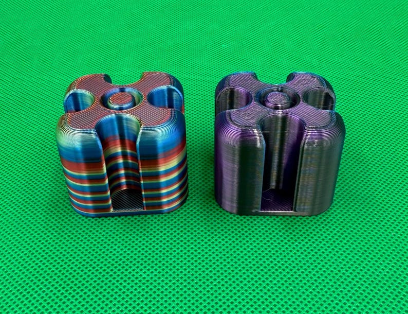 May include: Two 3D printed square-shaped objects with rounded corners and a central circular opening. One object features horizontal stripes of red, blue, and yellow, while the other has a dark, iridescent purple and blue finish.