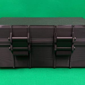 Puede incluir: Una caja de almacenamiento rectangular negra con una superficie texturizada y una tapa plana. La parte frontal del contenedor cuenta con dos mecanismos de cierre verticales que aseguran la tapa a la base, ofreciendo una soluci&oacute;n de almacenamiento robusta.