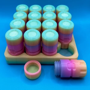 May include: A collection of small, cylindrical containers with screw-on lids, featuring a gradient color pattern transitioning from pink to orange to light blue. The containers are arranged in a tan tray with one empty slot, with one lid and one container placed separately in front.