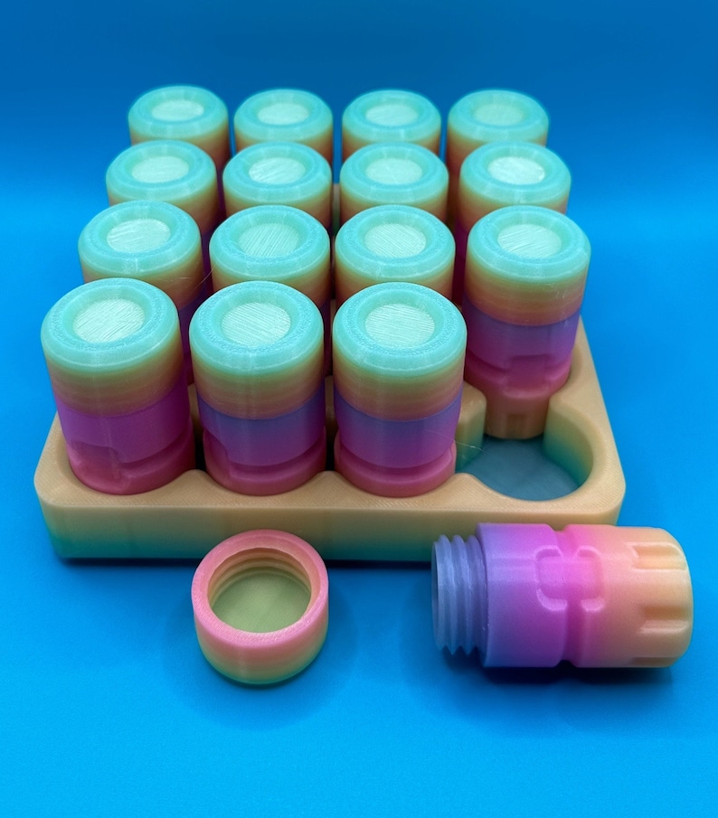 May include: A collection of small, cylindrical containers with screw-on lids, featuring a gradient color pattern transitioning from pink to orange to light blue. The containers are arranged in a tan tray with one empty slot, with one lid and one container placed separately in front.
