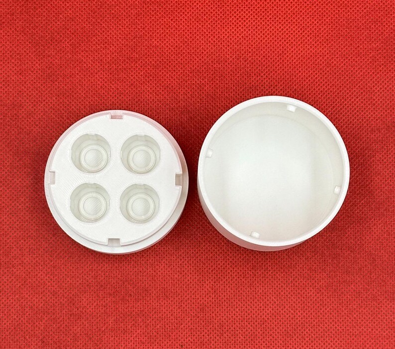Op de afbeelding: Een witte ronde kunststof container met een apart deksel voorzien van vier kleine, verzonken ronde vakjes. Het deksel ligt aan de linkerkant, terwijl de open, holle cilindrische basis aan de rechterkant staat.