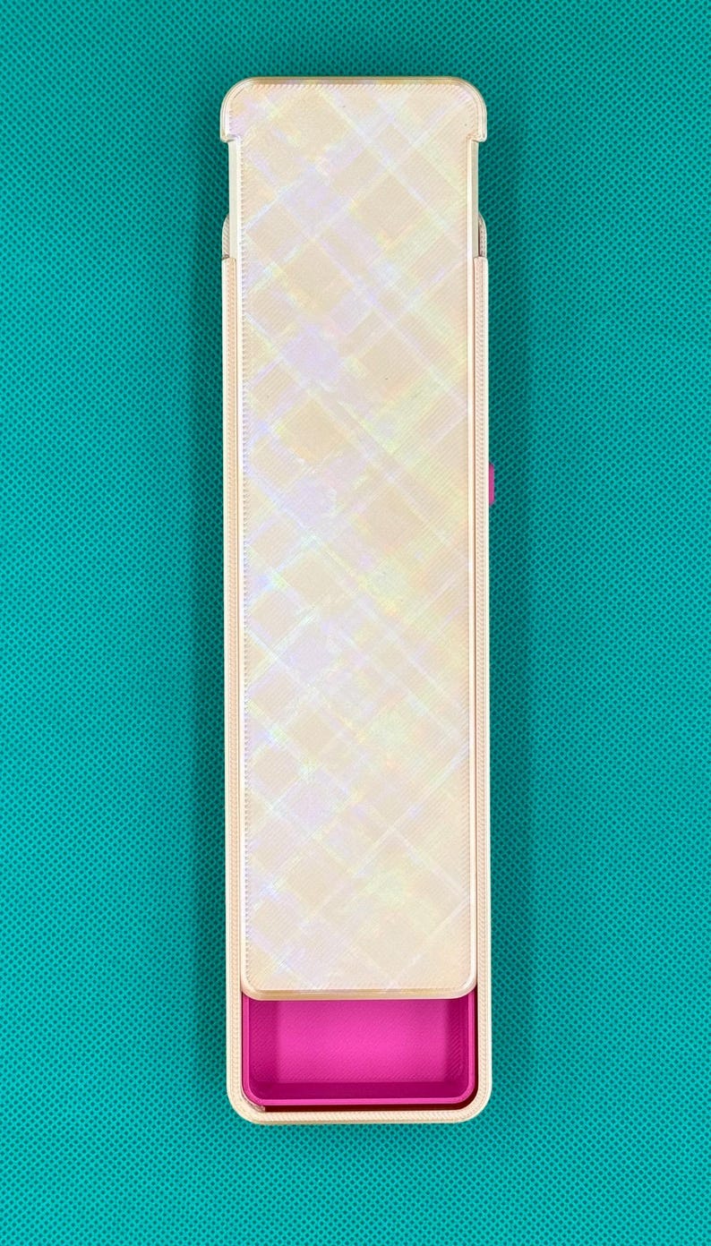 May include: A rectangular storage case with a sliding lid featuring a pearlescent, iridescent finish with a diagonal grid pattern. The base of the case is a solid, vibrant magenta pink color, visible at the bottom where the lid is partially open.