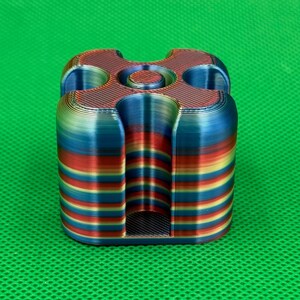 May include: A 3D printed square-shaped object with rounded corners and a central circular opening. The item features horizontal layers of gradient colors including red, blue, yellow, and green, with four deep vertical grooves on each side.