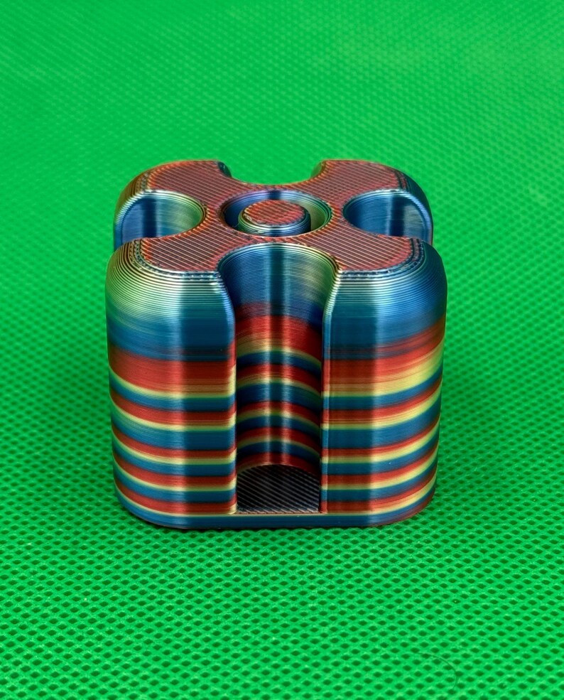 May include: A 3D printed square-shaped object with rounded corners and a central circular opening. The item features horizontal layers of gradient colors including red, blue, yellow, and green, with four deep vertical grooves on each side.