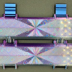 May include: Two iridescent purple and black rectangular boxes with a holographic heart pattern and a starburst design in the center. The boxes have blue plastic hinges on the top.