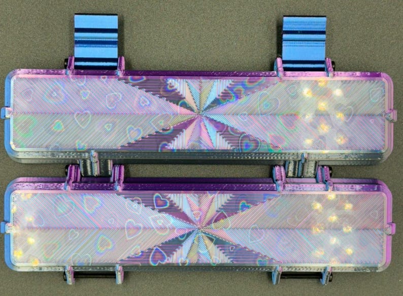 May include: Two iridescent purple and black rectangular boxes with a holographic heart pattern and a starburst design in the center. The boxes have blue plastic hinges on the top.