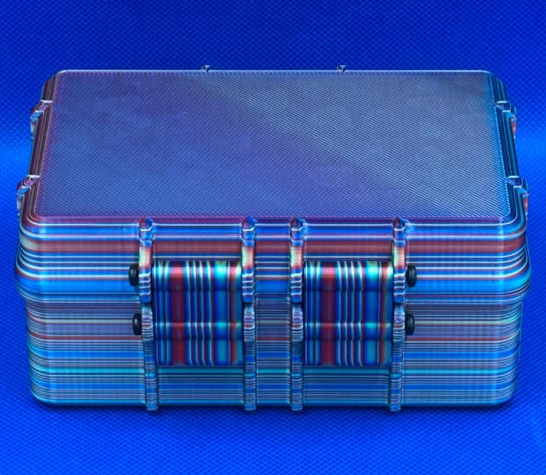 May include: A rectangular hard-shell storage case featuring a metallic, iridescent finish with horizontal bands of blue, purple, and red. The exterior has a textured, carbon-fiber-like pattern on the lid and two prominent front latches.