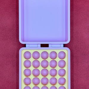 May include: A light purple rectangular storage case with two gold-colored metal latches at the top. The open bottom section contains a yellow insert with twenty-five circular indentations, each holding a small, round, light pink object.