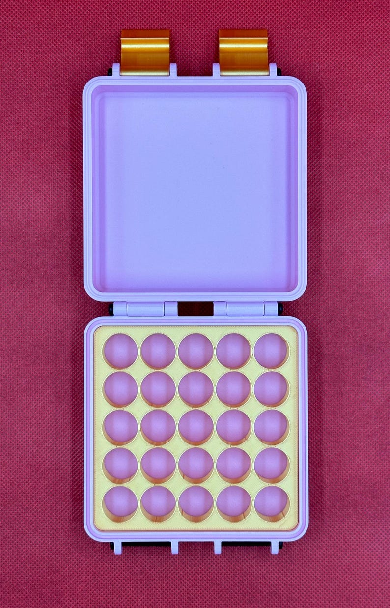 May include: A light purple rectangular storage case with two gold-colored metal latches at the top. The open bottom section contains a yellow insert with twenty-five circular indentations, each holding a small, round, light pink object.