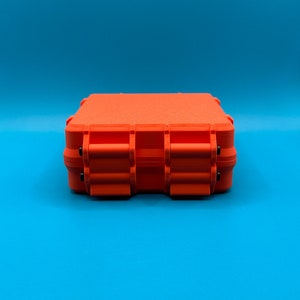 May include: A bright orange rectangular storage case with a textured surface and two prominent front latches. The case features a rigid, boxy construction with visible hinge details and small black pins securing the locking mechanisms.