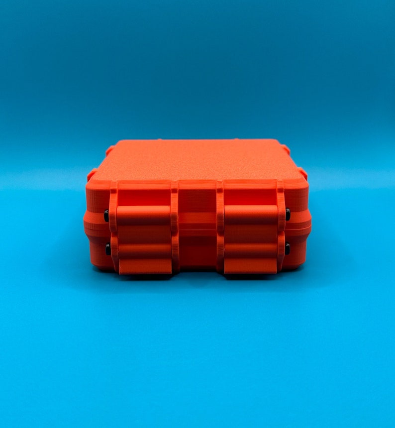 May include: A bright orange rectangular storage case with a textured surface and two prominent front latches. The case features a rigid, boxy construction with visible hinge details and small black pins securing the locking mechanisms.