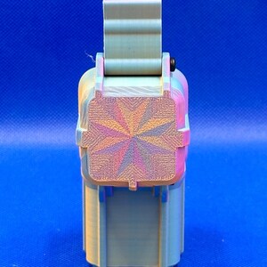 May include: A 3D printed rectangular storage container with a hinged lid. The exterior features a light green color with a subtle iridescent finish. The top of the lid displays a prominent starburst pattern with radiating lines in shades of pink, blue, and gold.