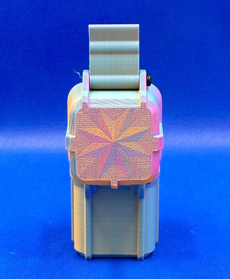 May include: A 3D printed rectangular storage container with a hinged lid. The exterior features a light green color with a subtle iridescent finish. The top of the lid displays a prominent starburst pattern with radiating lines in shades of pink, blue, and gold.