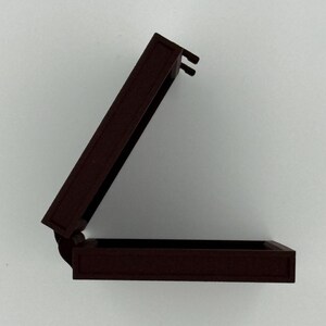 May include: A small, open, rectangular box with a hinged lid. The box is a dark reddish-brown color, with a matte finish. The lid is slightly ajar, revealing the interior of the box. The box appears to be made of wood or a similar material.
