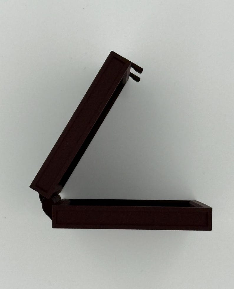 May include: A small, open, rectangular box with a hinged lid. The box is a dark reddish-brown color, with a matte finish. The lid is slightly ajar, revealing the interior of the box. The box appears to be made of wood or a similar material.