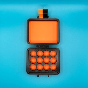 May include: A rectangular black storage case with an orange interior. The top lid is open, revealing a solid orange lining. The bottom section features a grid of twelve circular orange indentations. A black and orange latch is at the top.