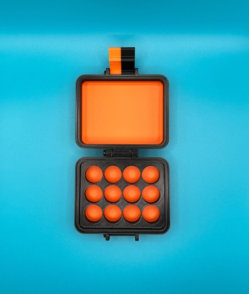 May include: A rectangular black storage case with an orange interior. The top lid is open, revealing a solid orange lining. The bottom section features a grid of twelve circular orange indentations. A black and orange latch is at the top.
