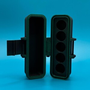 Puede incluir: Un estuche de almacenamiento rectangular con bisagras hecho de material verde oscuro y negro. El estuche est&aacute; abierto, revelando un compartimento hueco en el lado izquierdo y una bandeja con cinco ranuras circulares verticales a la derecha.