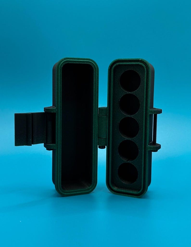 Puede incluir: Un estuche de almacenamiento rectangular con bisagras hecho de material verde oscuro y negro. El estuche est&aacute; abierto, revelando un compartimento hueco en el lado izquierdo y una bandeja con cinco ranuras circulares verticales a la derecha.