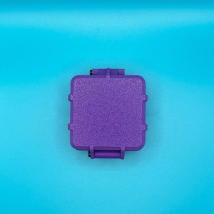 May include: A square, purple, textured storage case with rounded corners and a hinged lid. The case features a rugged, matte finish on the top surface and small black hardware components visible at the hinge points on the top and bottom edges.