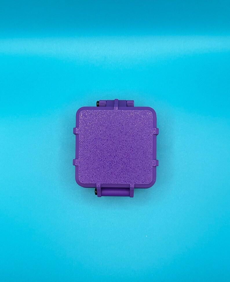 May include: A square, purple, textured storage case with rounded corners and a hinged lid. The case features a rugged, matte finish on the top surface and small black hardware components visible at the hinge points on the top and bottom edges.