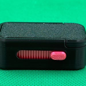 May include: A rectangular black plastic case featuring a textured top surface and a horizontal slot on the front. Inside the slot, a bright pink sliding mechanism with a ribbed grip allows for manual adjustment of the internal component.