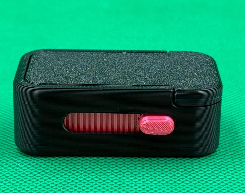May include: A rectangular black plastic case featuring a textured top surface and a horizontal slot on the front. Inside the slot, a bright pink sliding mechanism with a ribbed grip allows for manual adjustment of the internal component.