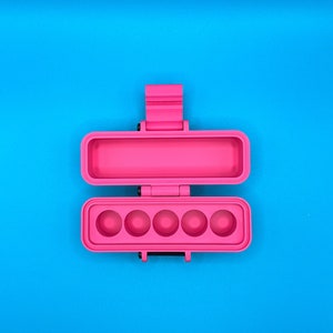 Puede incluir: Un estuche de almacenamiento rectangular de color rosa brillante con tapa abatible. El interior de la secci&oacute;n inferior presenta cinco hendiduras circulares en fila, mientras que la secci&oacute;n superior es un compartimento rectangular hueco.