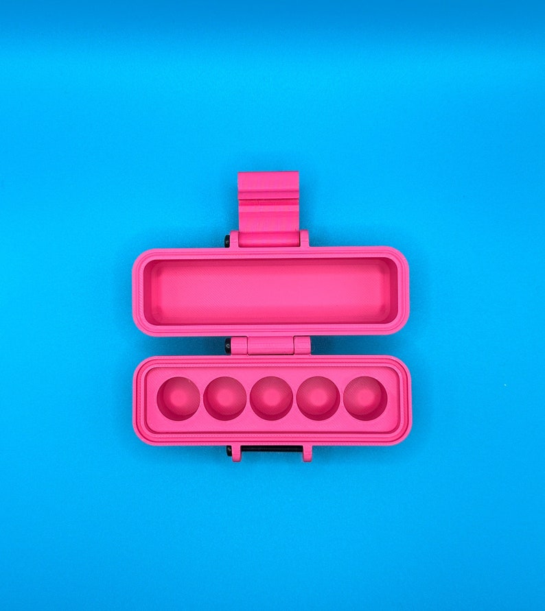 Puede incluir: Un estuche de almacenamiento rectangular de color rosa brillante con tapa abatible. El interior de la secci&oacute;n inferior presenta cinco hendiduras circulares en fila, mientras que la secci&oacute;n superior es un compartimento rectangular hueco.