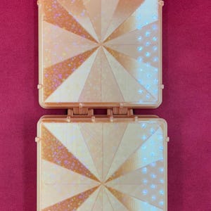 May include: A square, peach-colored plastic case with a hinged design. The front panels feature a radiating sunburst pattern with alternating sections of solid color and holographic star-shaped glitter accents. Two small hinges are visible at the top.