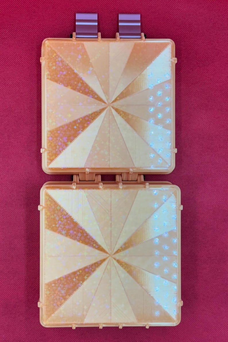 May include: A square, peach-colored plastic case with a hinged design. The front panels feature a radiating sunburst pattern with alternating sections of solid color and holographic star-shaped glitter accents. Two small hinges are visible at the top.