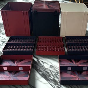 May include: A collection of storage containers and trays in various colors. The set includes a burgundy open-top box, a black box with a patterned lid, and a cream-colored box. Several trays with circular and rectangular compartments are also visible.