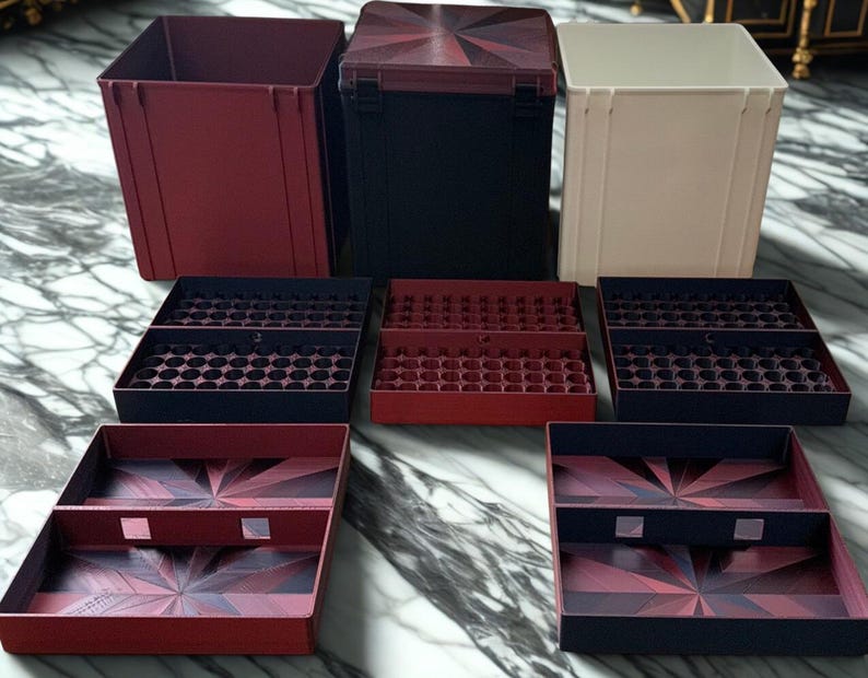 May include: A collection of storage containers and trays in various colors. The set includes a burgundy open-top box, a black box with a patterned lid, and a cream-colored box. Several trays with circular and rectangular compartments are also visible.