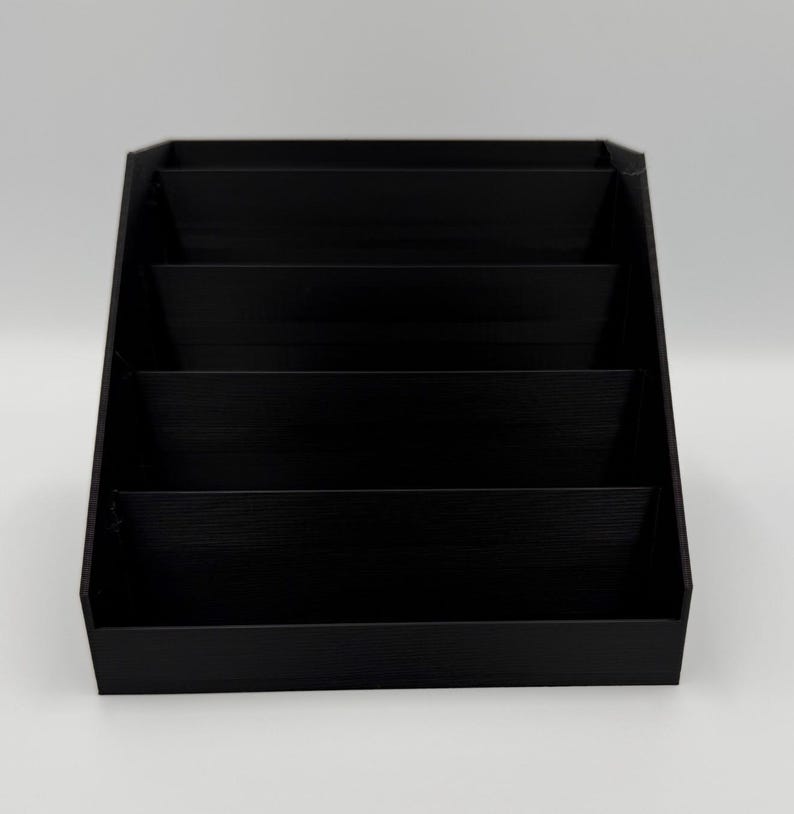 May include: A black, tiered display stand with four shelves. The stand is angled, with each shelf slightly recessed. The stand is made of a solid, matte black material, and is suitable for displaying small items.