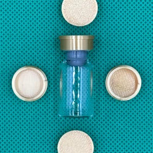 May include: A clear glass vial with a metallic gold-colored cap and a blue stopper, surrounded by four circular, light-colored, textured discs arranged in a cross pattern around the central glass container.