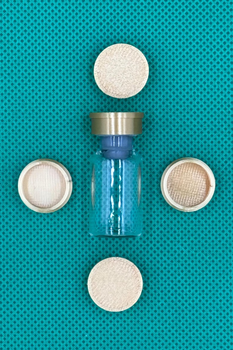May include: A clear glass vial with a metallic gold-colored cap and a blue stopper, surrounded by four circular, light-colored, textured discs arranged in a cross pattern around the central glass container.