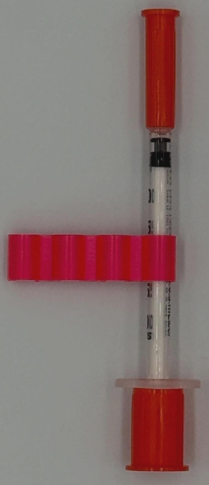 May include: A medical syringe with an orange cap and plunger, connected to a pink, ridged, rectangular component. The syringe is clear with black markings and a white interior. The components are set against a white background.