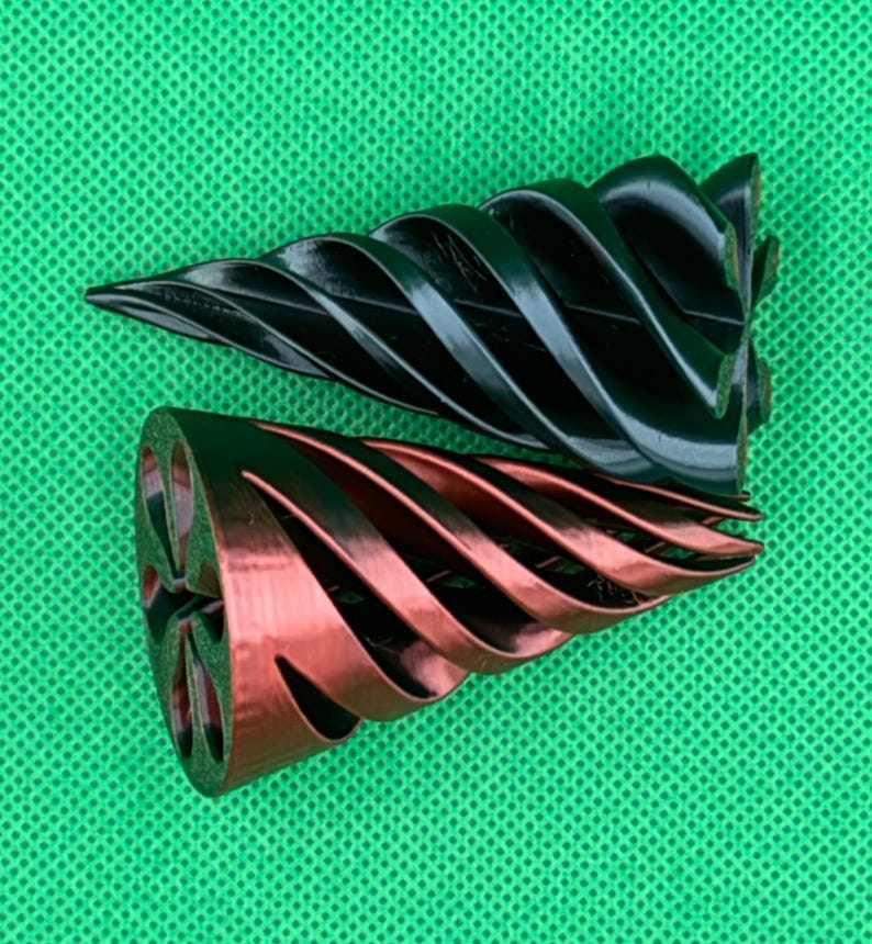 May include: Two conical objects with a spiral, ribbed design. One object is solid black, and the other is a metallic copper color with a dark interior. Both items feature a pointed tip and a wide, circular base with a six-petaled floral cutout pattern.