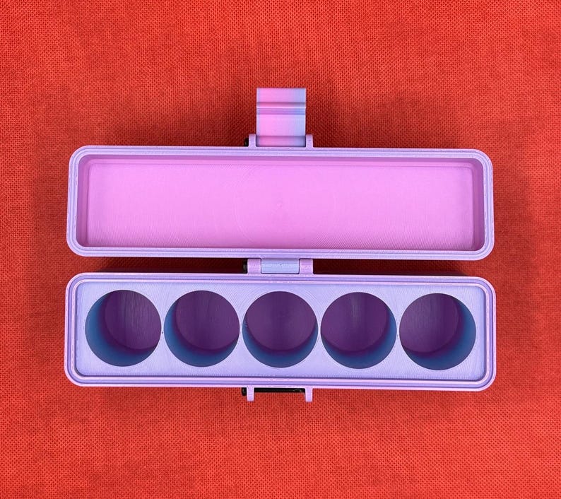 May include: A rectangular, hinged storage case featuring a light purple exterior and a gradient hinge mechanism. The interior of the base contains five circular, deep-set compartments, while the lid is open and empty.