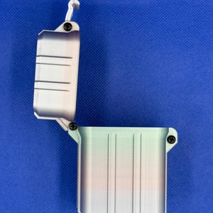 May include: An open, rectangular storage container with a hinged lid, featuring a vertical gradient colour pattern transitioning from light green to orange and purple. The box is secured with small black screws at the hinge and latch points.