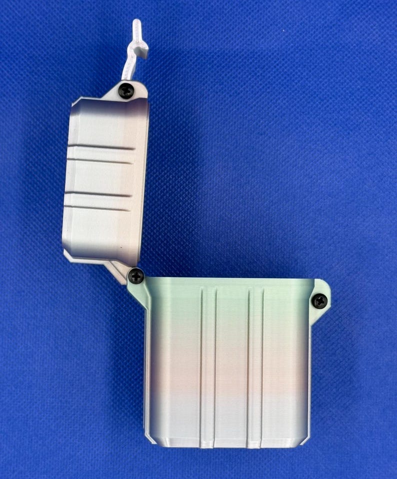 May include: An open, rectangular storage container with a hinged lid, featuring a vertical gradient colour pattern transitioning from light green to orange and purple. The box is secured with small black screws at the hinge and latch points.