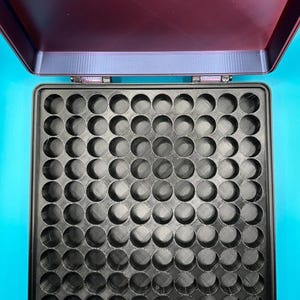 May include: A square black storage case with a hinged lid, featuring an interior grid of eighty-one circular compartments arranged in a nine-by-nine pattern. The lid is open, revealing a dark maroon interior surface with a subtle texture.