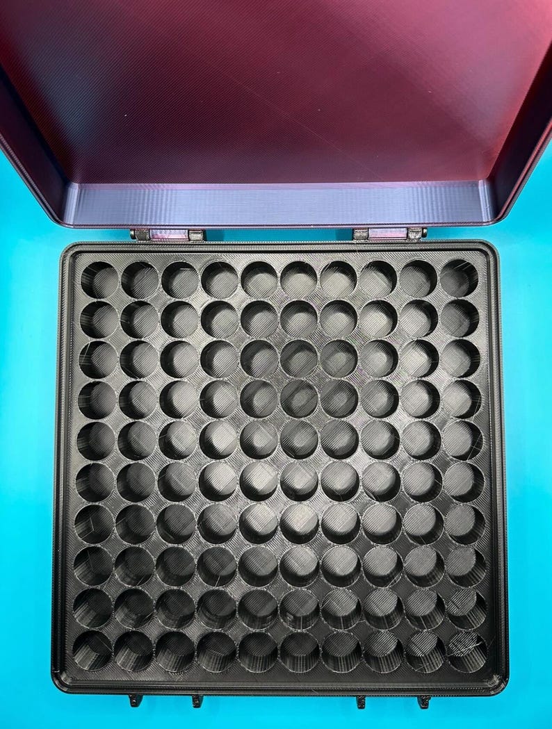 May include: A square black storage case with a hinged lid, featuring an interior grid of eighty-one circular compartments arranged in a nine-by-nine pattern. The lid is open, revealing a dark maroon interior surface with a subtle texture.