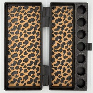 May include: A black rectangular hinged case featuring a tan interior with a black leopard print pattern. The right side of the case includes a column of seven circular cutouts designed for holding small cylindrical items securely.
