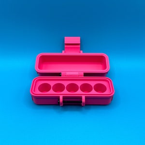 Puede incluir: Un estuche de almacenamiento rectangular de color rosa brillante con la tapa abierta. La base interior presenta cinco hendiduras circulares dispuestas en una fila horizontal. El estuche incluye una tapa con bisagras y un cierre con un pasador negro.
