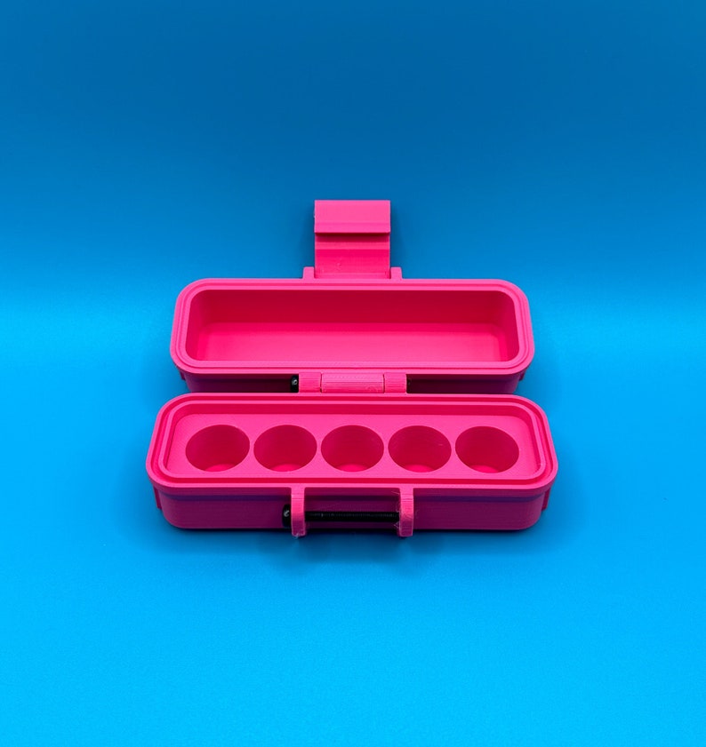 Puede incluir: Un estuche de almacenamiento rectangular de color rosa brillante con la tapa abierta. La base interior presenta cinco hendiduras circulares dispuestas en una fila horizontal. El estuche incluye una tapa con bisagras y un cierre con un pasador negro.
