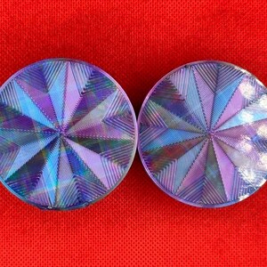 Op de afbeelding: Twee ronde oorplugs met een holografische, iriserende afwerking die verschuift tussen tinten paars, blauw en zilver. Het oppervlak van elke plug toont een geometrisch, zonnestraalachtig patroon van fijne, ge&euml;tste lijnen.