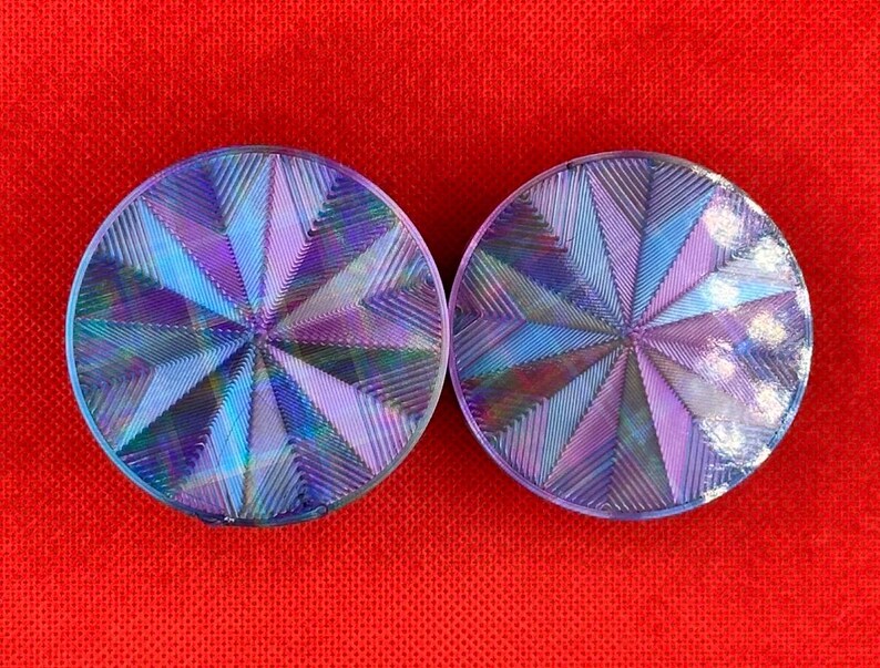 Op de afbeelding: Twee ronde oorplugs met een holografische, iriserende afwerking die verschuift tussen tinten paars, blauw en zilver. Het oppervlak van elke plug toont een geometrisch, zonnestraalachtig patroon van fijne, ge&euml;tste lijnen.