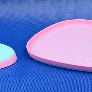 May include: Two pastel-colored, oval-shaped serving trays. The larger tray is pink with a light blue rim. The smaller tray is light blue with a pink rim.